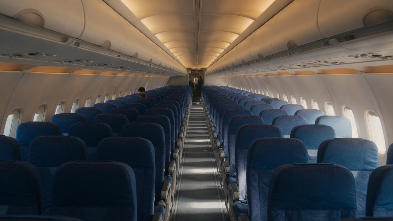 The empty cabin of an airliner seen down the centre aisle, rows of unoccupied deep blue seats with the overhead bins closed and one raised window shade letting in daylight