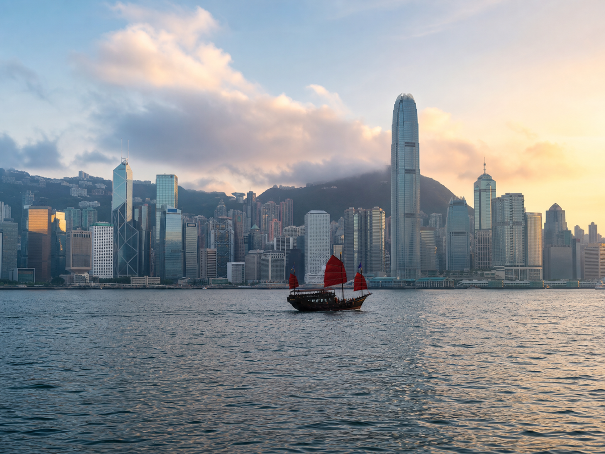 The Hong Kong skyline across Victoria Harbour at dawn, with a red-sailed junk on the water