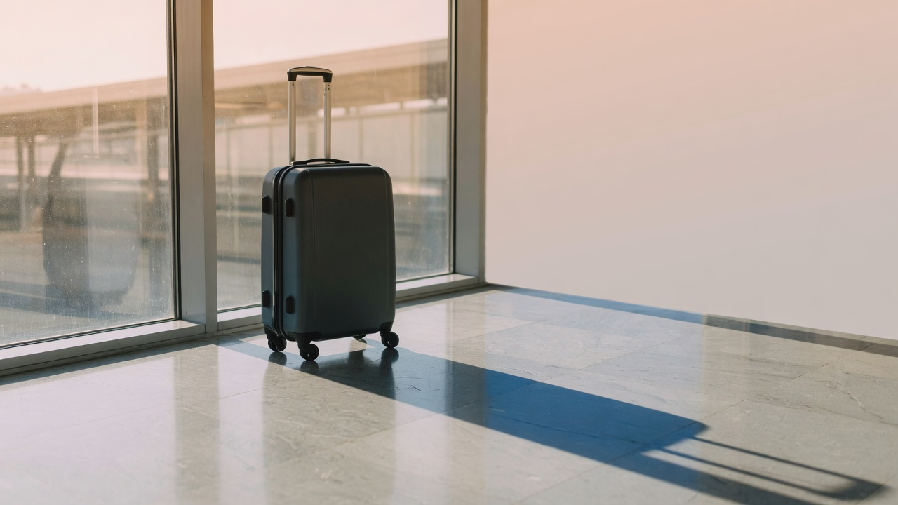 A single cabin-sized hard-shell roller case standing alone on a polished terminal floor beside a tall window, its handle extended and its shadow long in the morning light