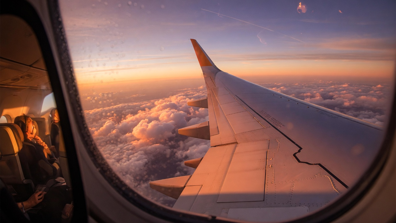 The wing of an airliner seen through a cabin window above a layer of cloud at sunset, with passengers seated in the row behind