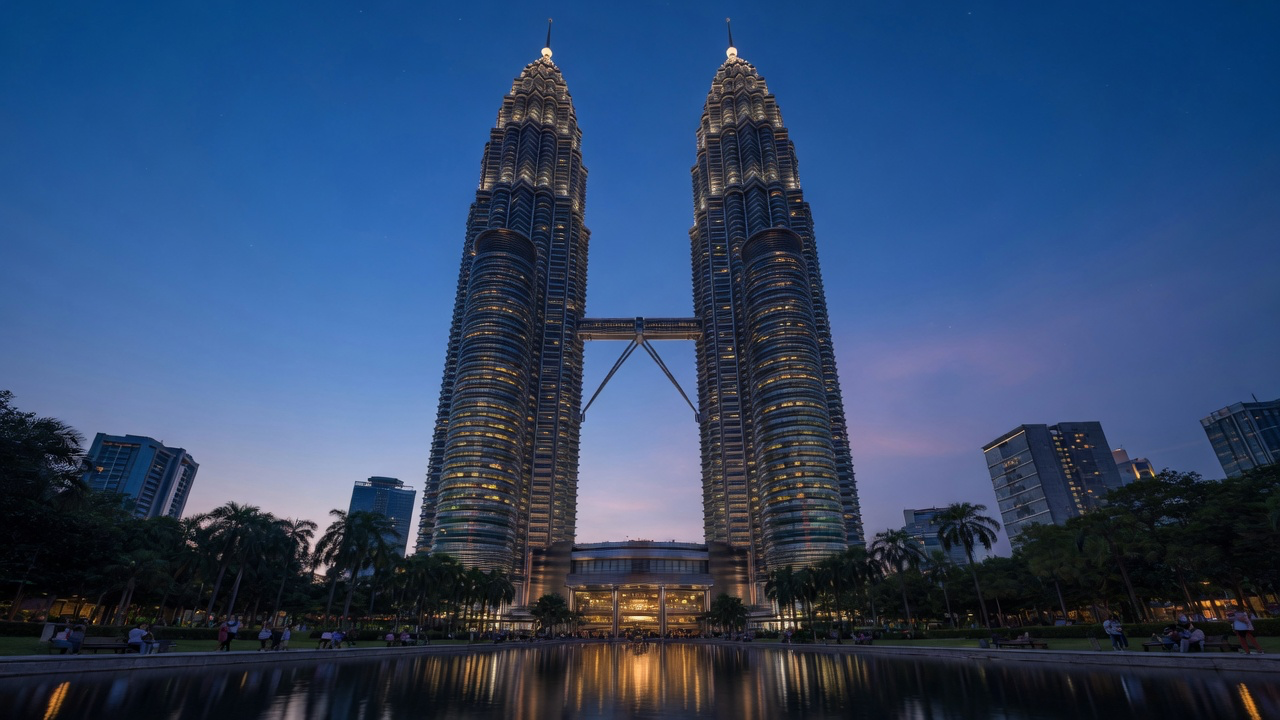 The Petronas Twin Towers in Kuala Lumpur at blue hour, lit from within and reflected in the still lake of KLCC Park, with palm trees and lower city towers around them