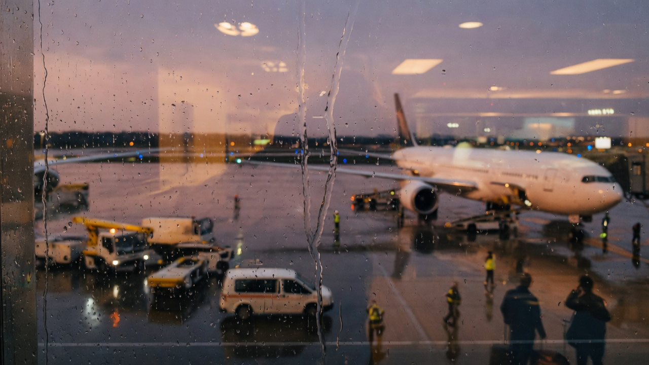 A rain-streaked airport terminal window at dusk, water running down the glass with the blurred shape of a parked aircraft and ground vehicles on the wet apron beyond