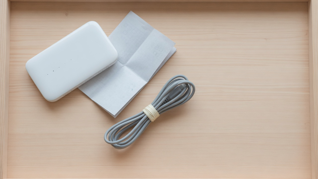 A small white pocket wifi router, a folded paper leaflet and a grey cable tied with a paper band on a pale hinoki wood tray in soft shadowless light
