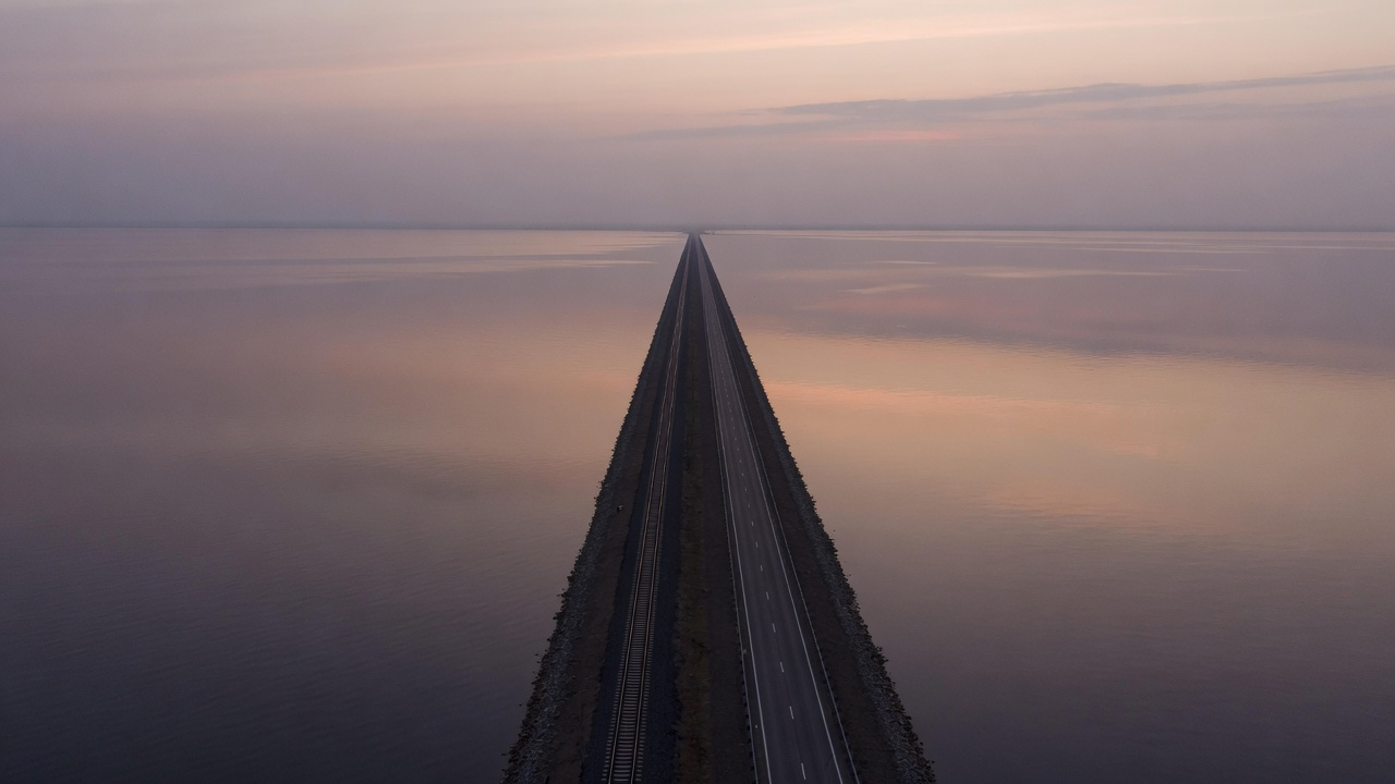 An aerial view at dusk of a long low causeway carrying a road and a parallel rail line dead straight across wide calm water, the horizon lost in haze and pale reflections on the surface either side