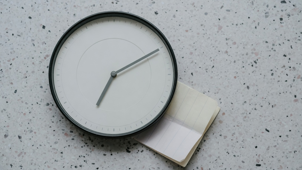 A round wall clock with no numerals and plain grey hands, resting on pale speckled terrazzo beside a small notepad