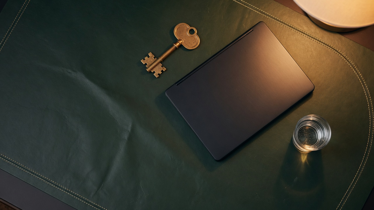 A closed slim laptop, a large brass key and a glass of water on a dark green stitched leather desk pad under warm lamplight at night