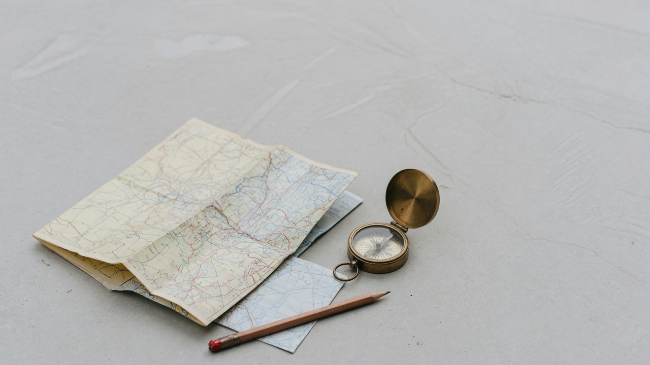 A folded paper road map, a brass lensatic compass and a short pencil on pale concrete in flat overcast light