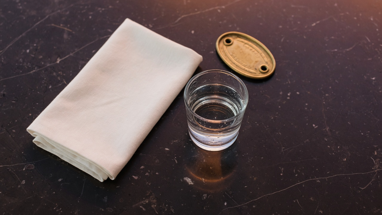 A folded cream linen napkin, a glass of water and a small brass token on a dark marble table with a soft sheen