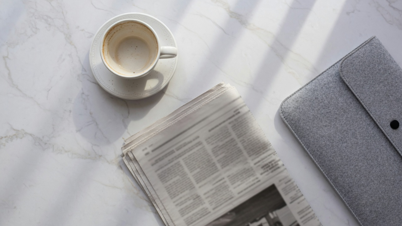 A white cappuccino cup on a saucer, a folded newspaper and a grey felt laptop sleeve on pale veined marble in bright diffused daylight