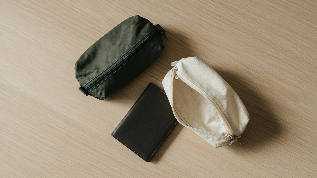 An olive ripstop pouch lying closed beside an open cream canvas pouch, with a dark brown leather wallet between them, on pale oak in soft daylight
