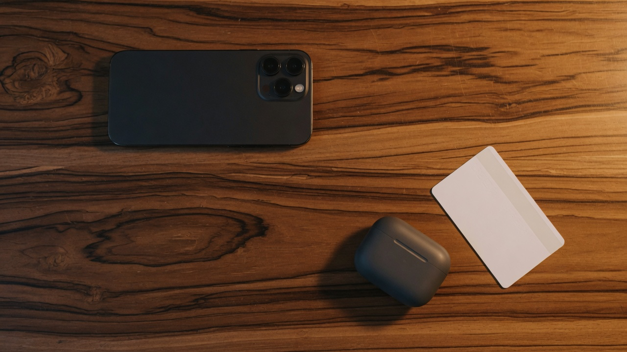 A dark phone lying face down showing its camera bump, a matte earbuds case and a plain white hotel key card on richly grained teak in warm evening light