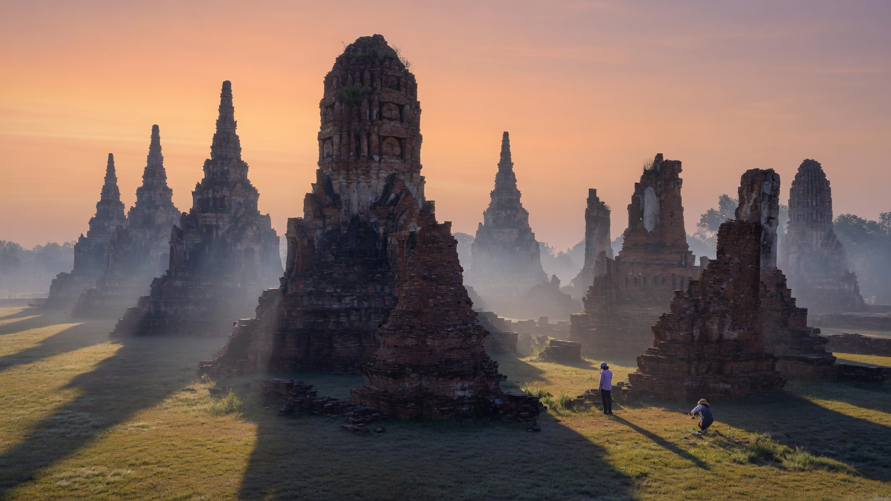 The brick prangs and ruined stupas of Ayutthaya at sunrise, long shadows across the grass and mist drifting between the ancient walls