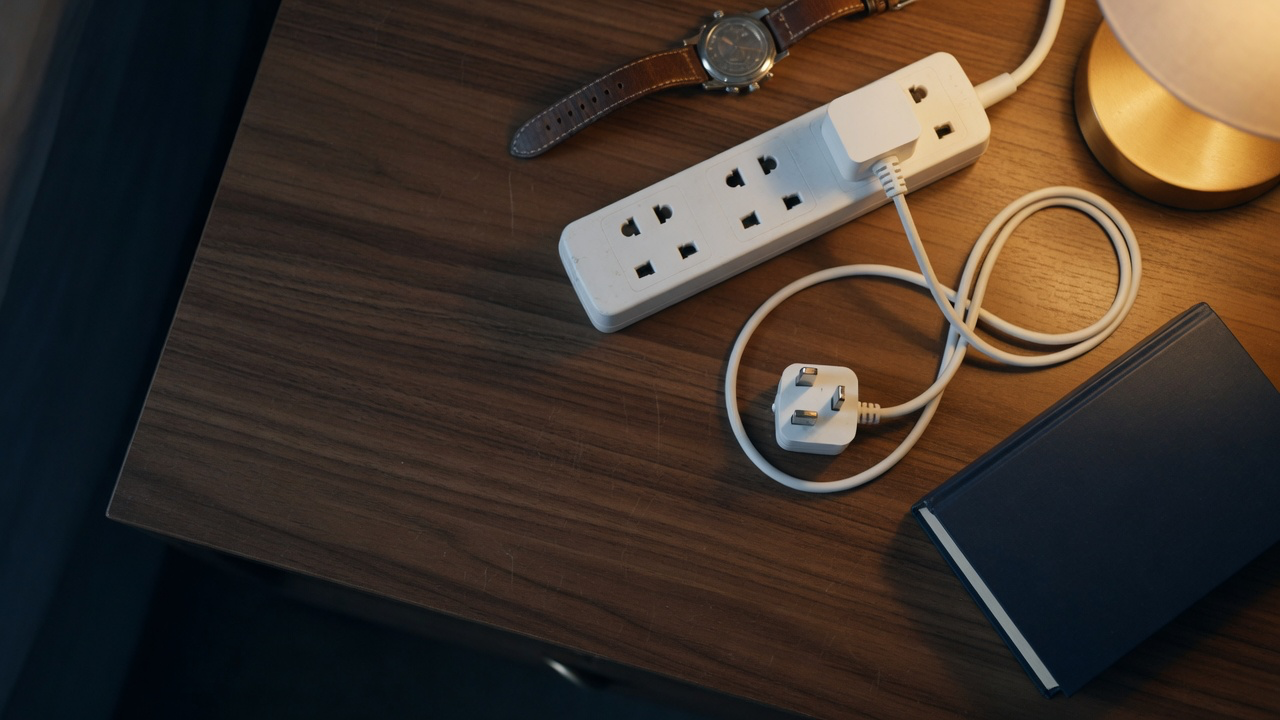A white Type G power strip with a plugged-in cable, a loose three-pin plug, a leather-strapped watch and a closed book on a dark walnut nightstand under a bedside lamp