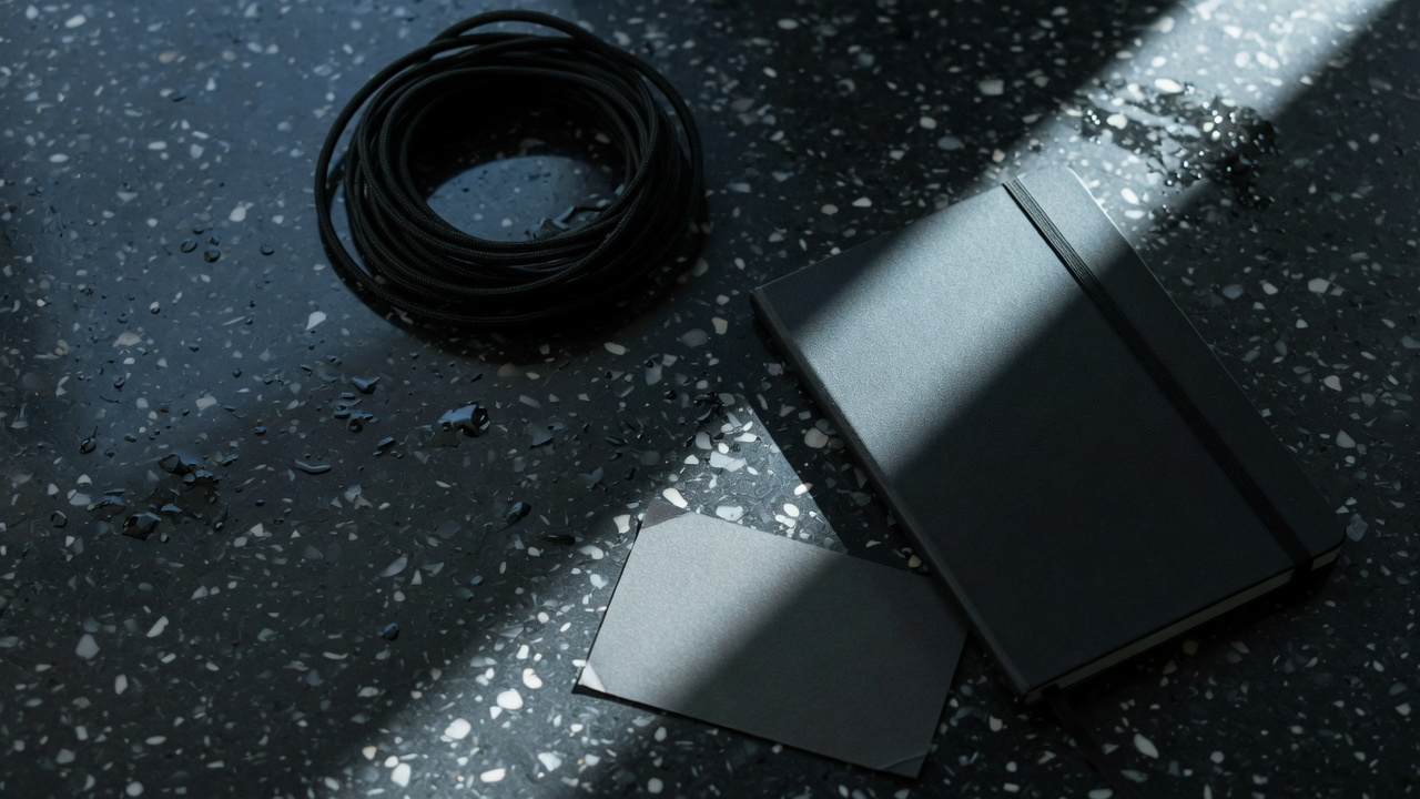 A tightly coiled black cable, a closed black notebook and a dark grey card on wet-look speckled terrazzo, cut by a single hard slash of cool light